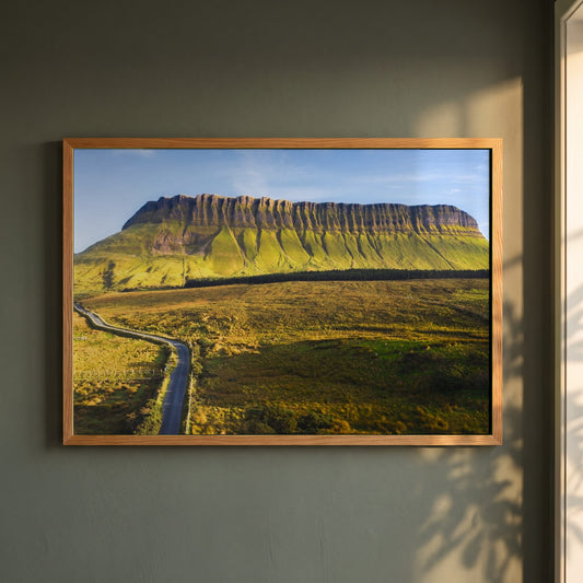 Benbulben, Sligo framed print in a natural oak frame on a dark green wall beside a window, dappled plant shadow