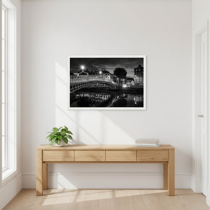 Black and white Ha’penny Bridge night photograph with city lights reflecting in the River Liffey, framed in a white wooden frame, 30x45 cm. Bright hallway with white walls and wooden console table, 30x45 cm Ha’penny Bridge black and white print in white frame hanging above, natural daylight, potted plant and books below, airy Dublin apartment vibe