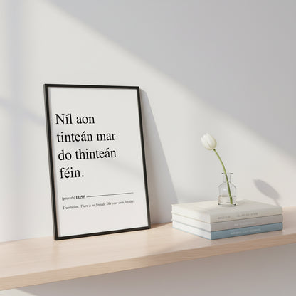 Níl aon tinteán mar do thinteán féin – Irish Gaelic “No Place Like Home” Print Framed A4 Irish proverb print leaning on a shelf against gallery-white wall, stacked art books, small bud vase, natural daylight.