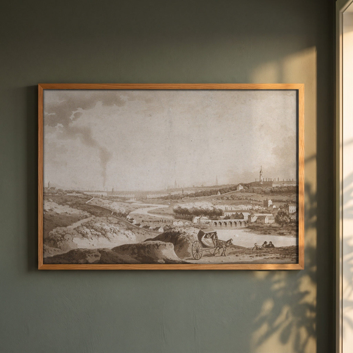 Dublin and Islandbridge from the Magazine Hill (1792) framed print in a natural oak frame on a dark green wall beside a window, dappled plant shadow