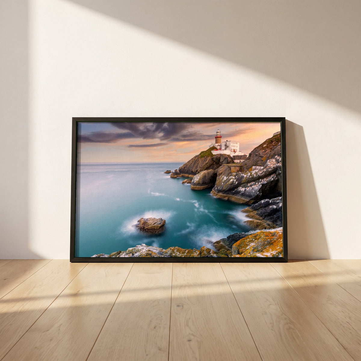 Baily Lighthouse Sunset, Howth Head framed print in a black frame leaning against a white plaster wall on oak floorboards in morning light