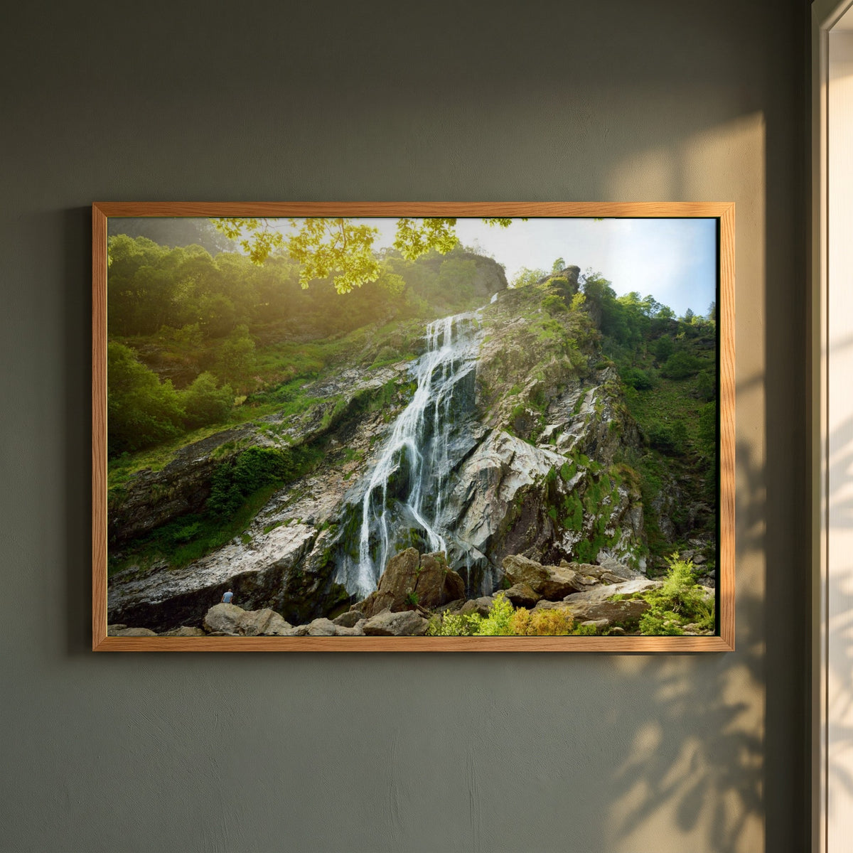 Powerscourt Waterfall Wicklow framed print in a natural oak frame on a dark green wall beside a window, dappled plant shadow