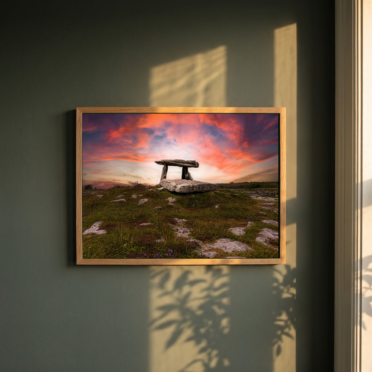 Poulnabrone Dolmen framed print in a natural oak frame on a dark green wall beside a window, dappled plant shadow