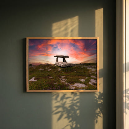 Poulnabrone Dolmen framed print in a natural oak frame on a dark green wall beside a window, dappled plant shadow