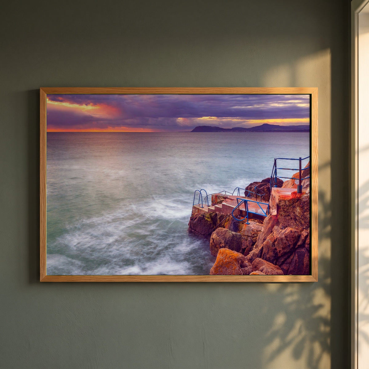 Vico Swimming Dalkey framed print in a natural oak frame on a dark green wall beside a window, dappled plant shadow